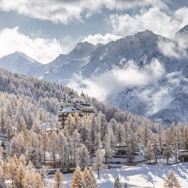 A serene winter scene featuring a large white building in a snowy forest, with mountains and a partly cloudy blue sky.