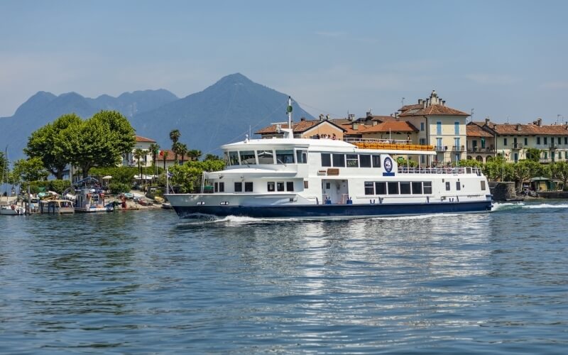 A white boat with a blue bottom navigates calm waters, surrounded by misty mountains and charming houses with red roofs.