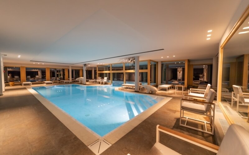 Serene indoor pool area with light blue water, beige tiles, white lounge chairs, and soft ambient lighting.