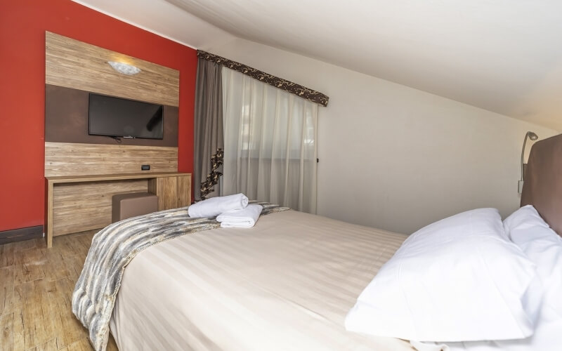 Cozy hotel room with a bed, desk, TV, and window, featuring a red accent wall and light wood flooring.