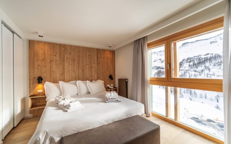 Camera serena con vista sulle montagne, letto bianco, pannelli in legno chiaro e finestre con tende grigie.