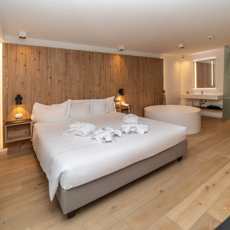 Modern hotel room with a large bed, nightstands, round bathtub, and bathroom sink, featuring light wood paneling and hardwood floor.