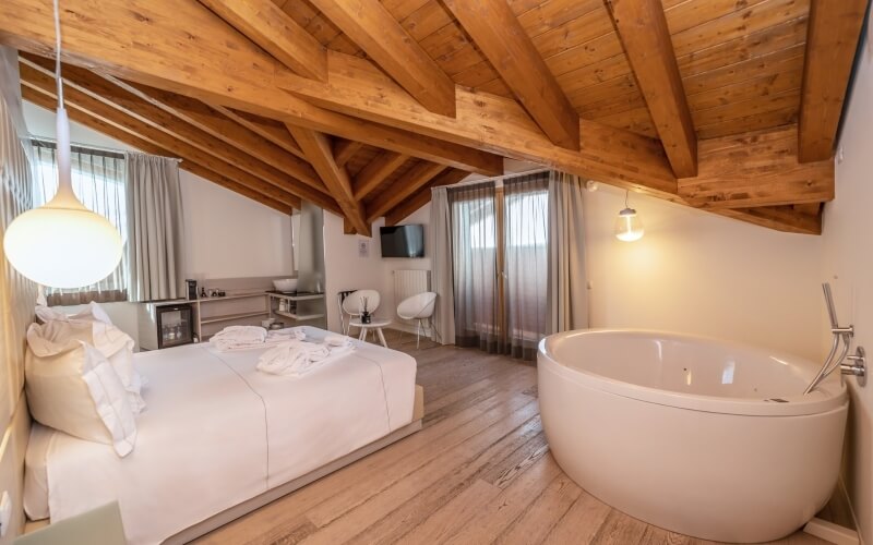 Minimalist hotel room with a white bed, large bathtub, wooden ceiling, and a small table by the window, exuding calmness.