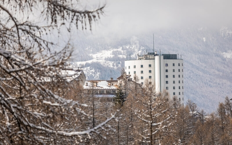 Scena invernale serena con un edificio bianco e montagne innevate sullo sfondo, alberi coperti di neve in primo piano.