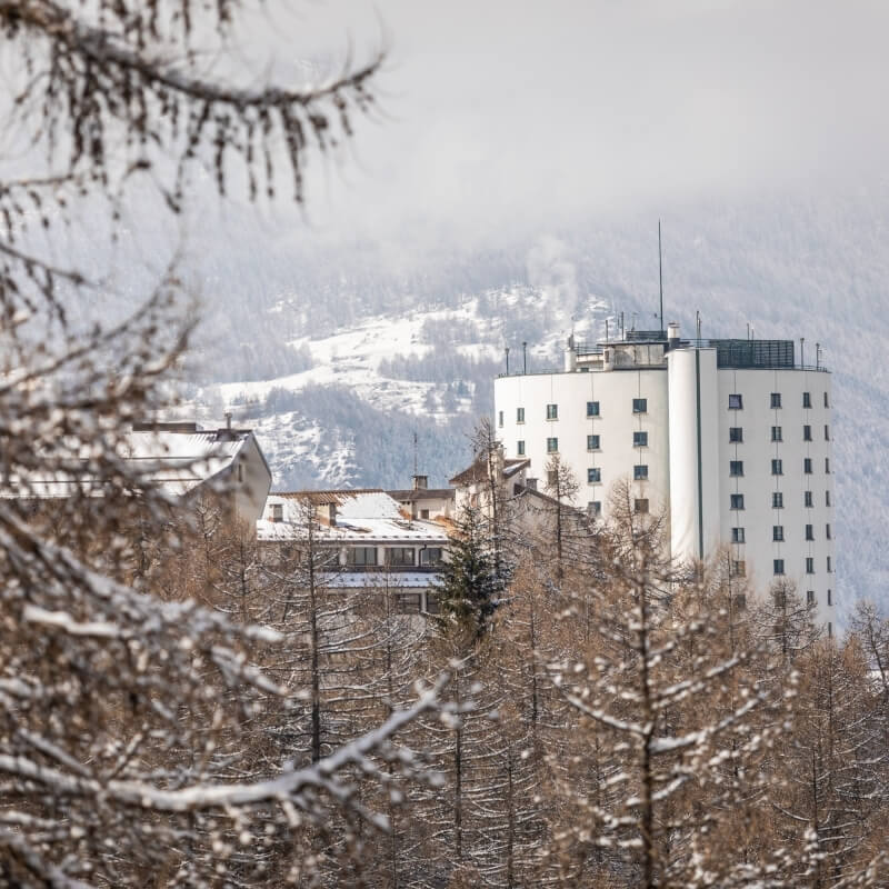 Serene winter scene with a large white building, bare trees, and snow-covered mountains under an overcast sky.