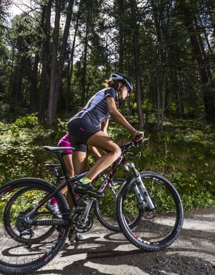 Two women in cycling outfits ride mountain bikes on a dirt path through a lush forest under a sunny sky.