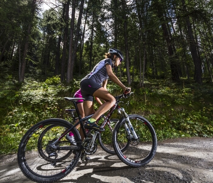 Two women in helmets ride mountain bikes on a forest path, surrounded by greenery and a glimpse of blue sky above.