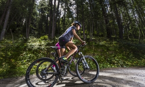 Two women biking on a dirt road in a wooded area, one in purple and the other in black, enjoying a sunny day.