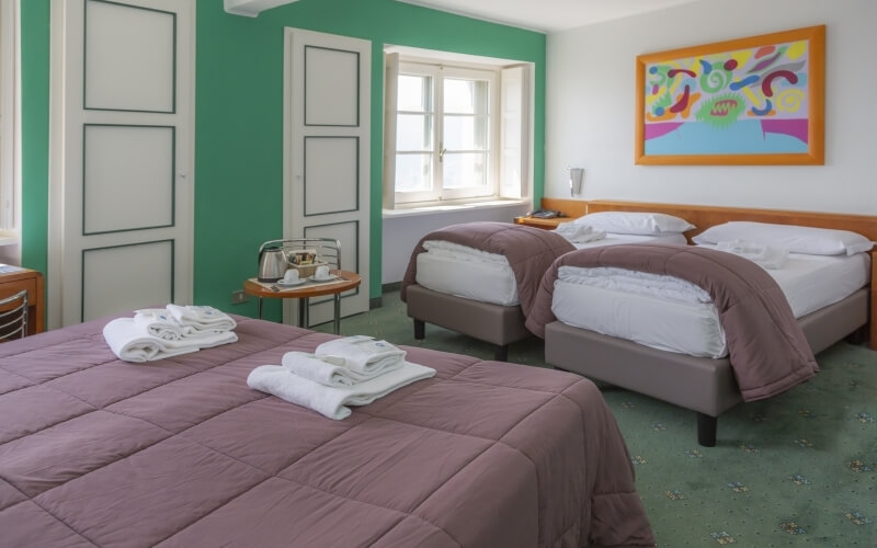 Hotel room with two beds, a round table, and a window; green and white walls, colorful artwork, and organized decor.