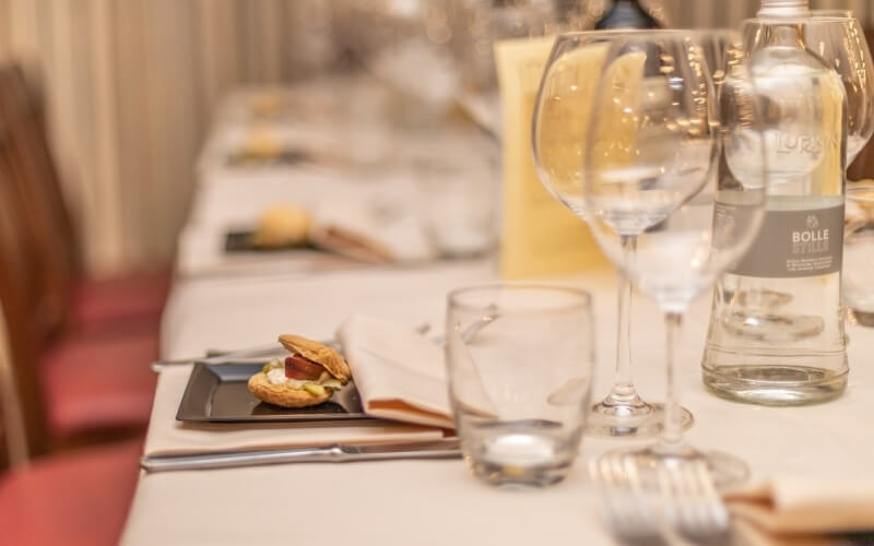 Formally set table with an open-faced sandwich, wine glasses, and a blurred second table, on a crimson carpet.