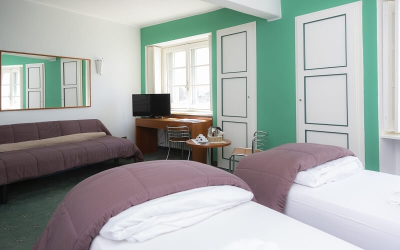 Hotel room with two beds, a desk, a daybed, and a window, featuring teal walls and a floral-patterned carpet.