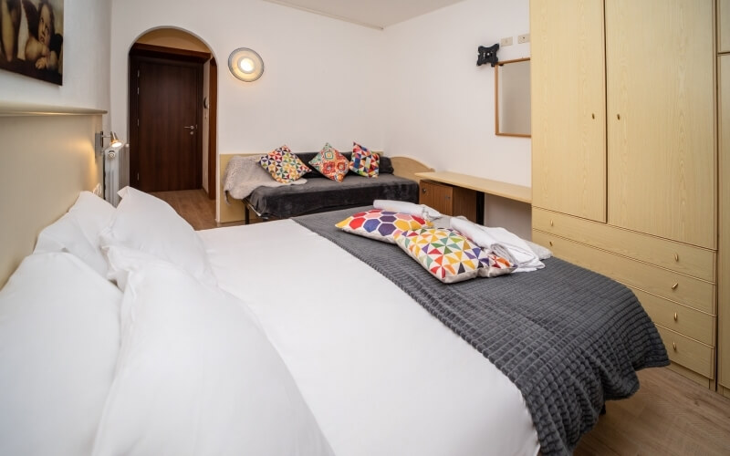 Camera d'hotel con due letti, uno rifatto con cuscini colorati, divano, scrivania e specchio su pareti bianche.