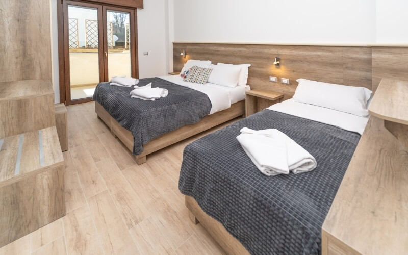 Serene bedroom with two beds, white linens, dark gray blankets, colorful pillow, and sliding glass door to a balcony.