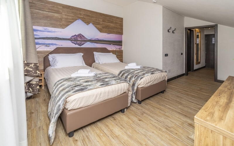 Cozy hotel room with two beds, mountain painting on wooden wall, hallway, dresser, and upholstered chair.