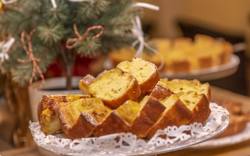 A plate of sliced yellow cake with a brown crust in front of a small decorated Christmas tree and another whole cake.