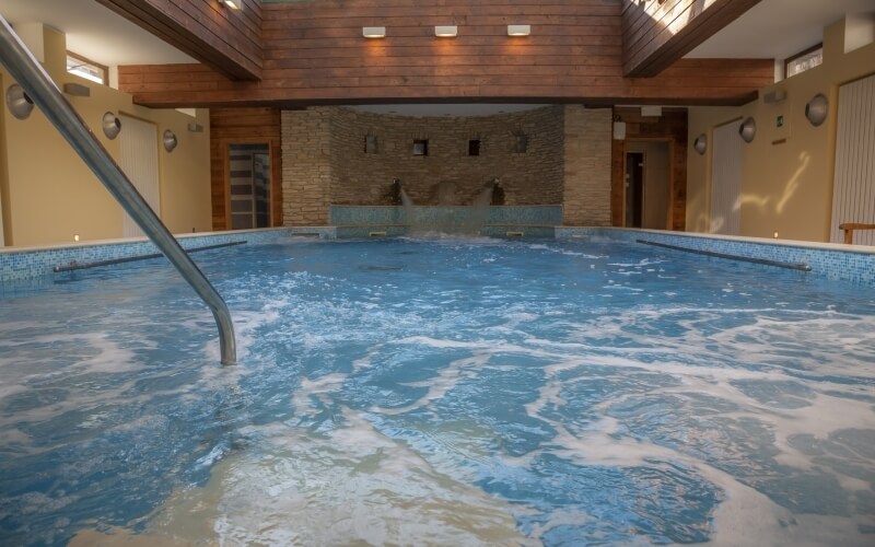 Indoor pool scene with light blue tiles, calm water, stone wall, wooden beam, and warm yellow walls, creating a serene atmosphere.