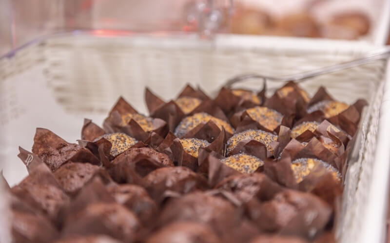 Close-up of uncooked brownies in paper liners, topped with yellow sauce and poppy seeds, in a white wicker basket.