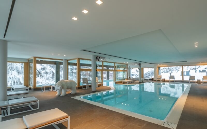 Area piscina interna moderna con acqua blu calma, sedie a sdraio bianche e grandi finestre che mostrano un paesaggio innevato.