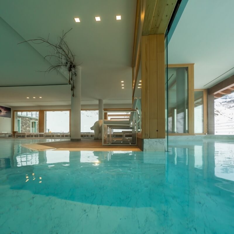 Piscina interna con acqua blu, pareti in legno e vetro, soffitto bianco con luci e area salotto sullo sfondo.