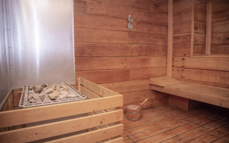Interno di una sauna in legno con panchina, griglia di metallo con rocce e un secchio di legno, atmosfera accogliente.