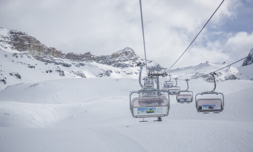 Ski lift in operation over snowy slopes, with a majestic snow-capped mountain range and cloudy sky in the background.