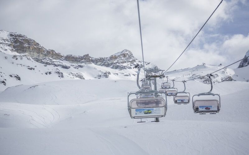 Ski lift with five transparent chairs ascends a snowy mountain; two chairs occupied by skiers, three empty, overcast sky.