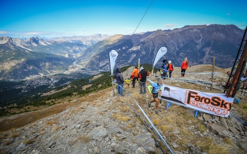 Gruppo di persone su una collina rocciosa con un cartello "Faro Sky Challenge" e bandiere, in un paesaggio montano.