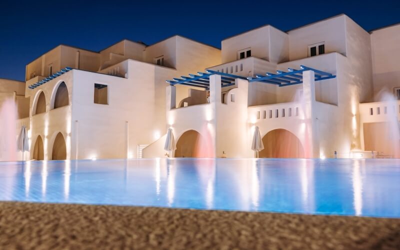Serene outdoor setting with a large blue pool reflecting lights from a multi-level white building under a dark blue sky.