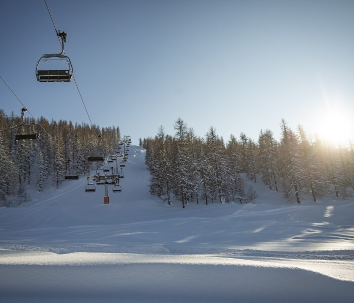 A ski lift with occupied chairs above a snowy slope, surrounded by trees and a bright sun under a clear blue sky.