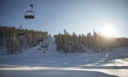 Ski lift with empty chairs ascends a snowy mountain under a bright blue sky and shining sun, creating a serene winter scene.