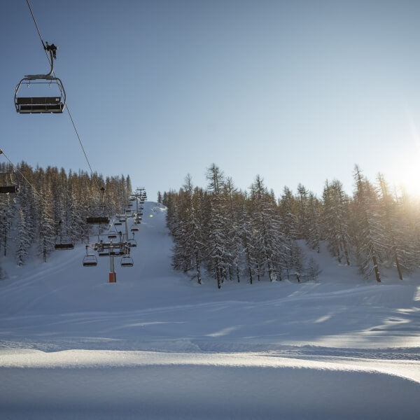 Serene snowy landscape with a ski lift, empty chairs, sun rising over trees, casting a warm glow on the snow.