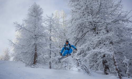 Snowboarder in a blue snowsuit airborne over snow-covered hills, with snow-dusted trees and a gray sky in the background.