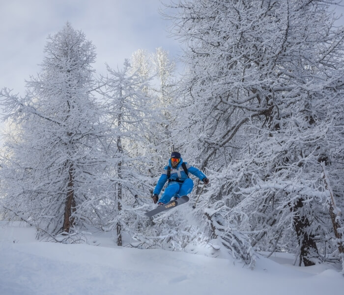Snowboarder in mid-air, wearing a blue and gray snowsuit, surrounded by snow-covered trees in a winter wonderland.