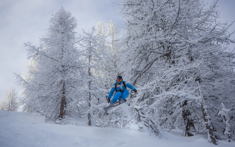 A snowboarder in a blue suit and black helmet jumps mid-air in a snowy landscape surrounded by snow-covered trees.