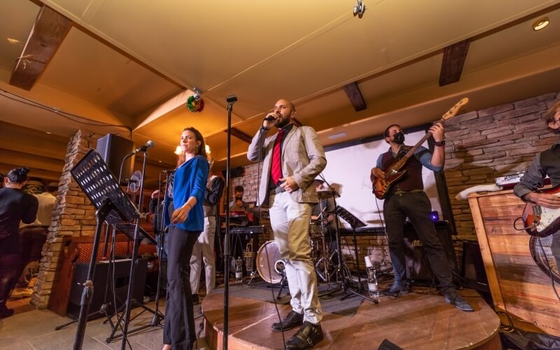 A band performs on stage with a lead singer in a white suit, flanked by a female vocalist and two guitarists, with a drummer behind.