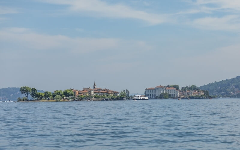 Scenic lakeside view with a charming town on a peninsula, calm blue water, trees, and a backdrop of hills under a light blue sky.