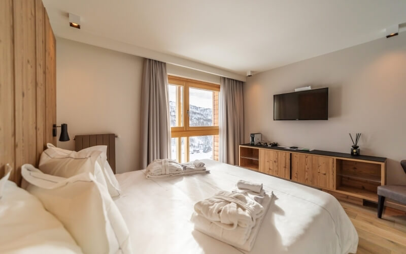 Camera d'albergo serena con letto bianco, TV a schermo piatto, luce naturale e tende grigie elegantemente tirate indietro.