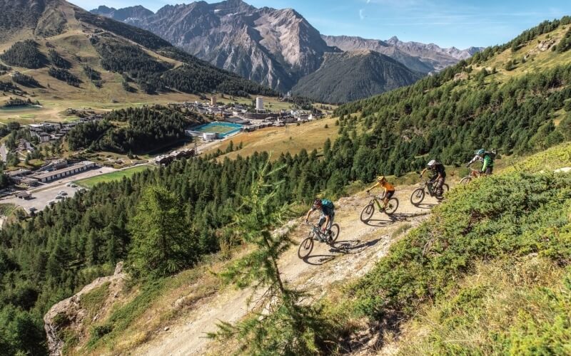 Mountain bikers ride on a dirt path through lush greenery, with hills and mountains in the background under a clear blue sky.