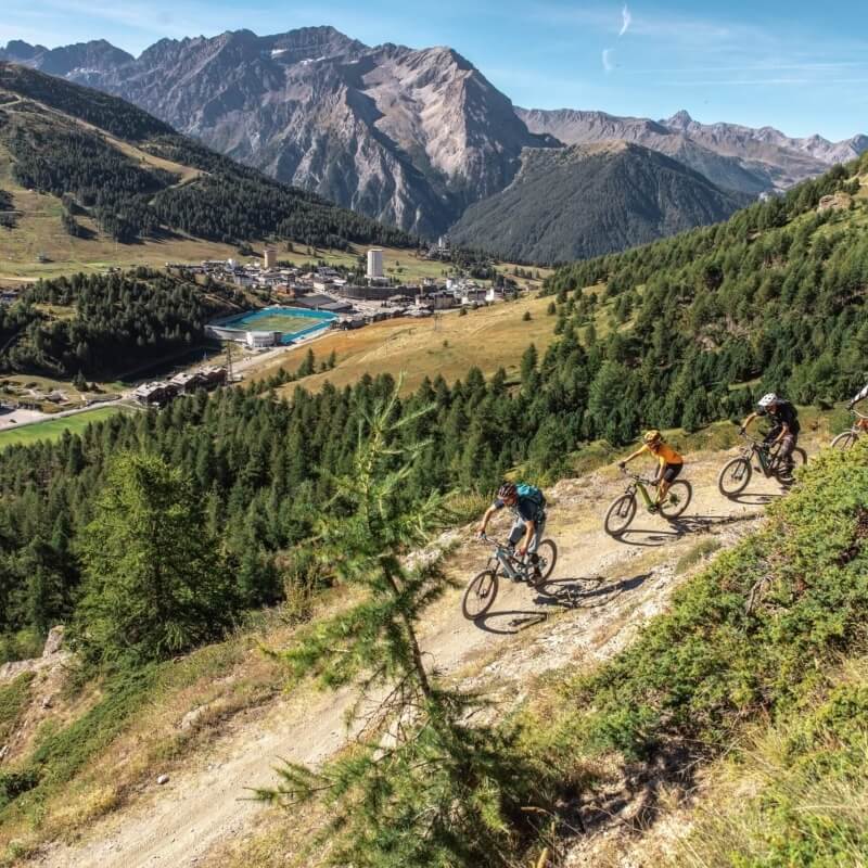 A group of four cyclists rides down a dirt path in a serene mountainous landscape under a clear blue sky.