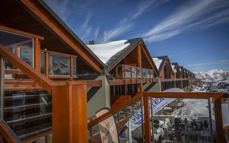 Ski resort with wooden buildings, a large snow-covered structure, and a blue sky, featuring distant snow-capped mountains.