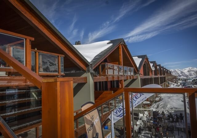 Modern building with sloping roof and wooden accents, set against snowy mountains, featuring a glass wall and winter sports vibe.