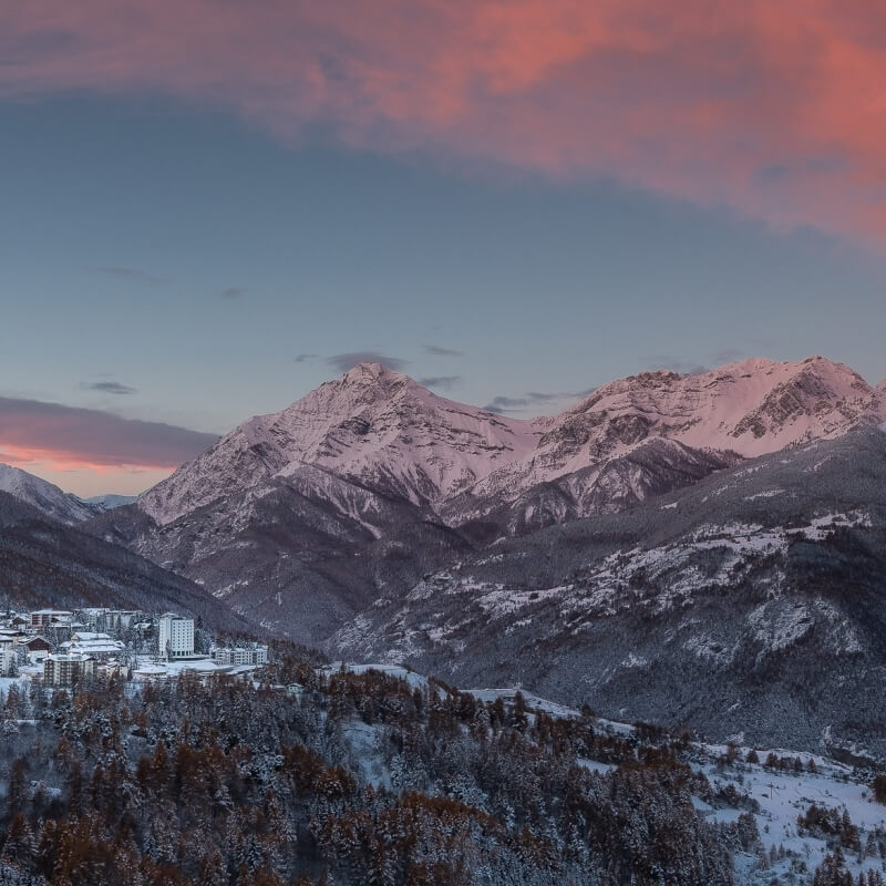 Snow-covered mountain range at sunset with a quaint village below and evergreen trees, creating a serene winter scene.