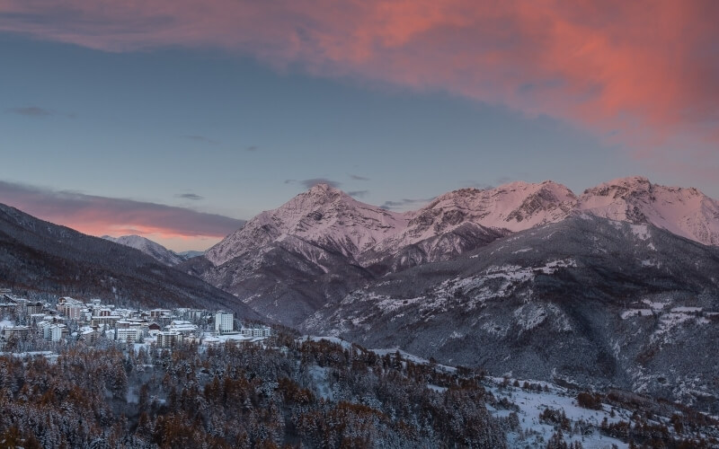 Catena montuosa innevata con un paese alla base, cielo blu che sfuma nel rosa, alberi imbiancati in primo piano.