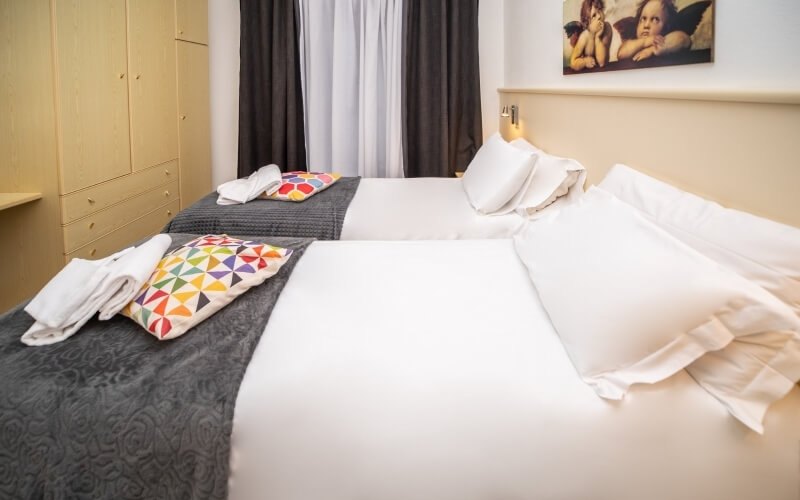 Camera d'hotel compatta con due letti, biancheria bianca, coperte grigie, cuscini colorati e decorazioni murali.