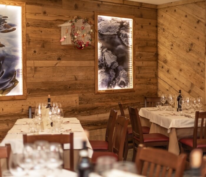 Rustic dining room with wooden walls, two long tables set for a meal, windows, framed nature pictures, and a birdcage.