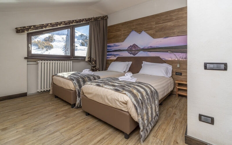 Hotel room with two beds, landscape painting, snowy view from window, modern decor, and warm ambiance.