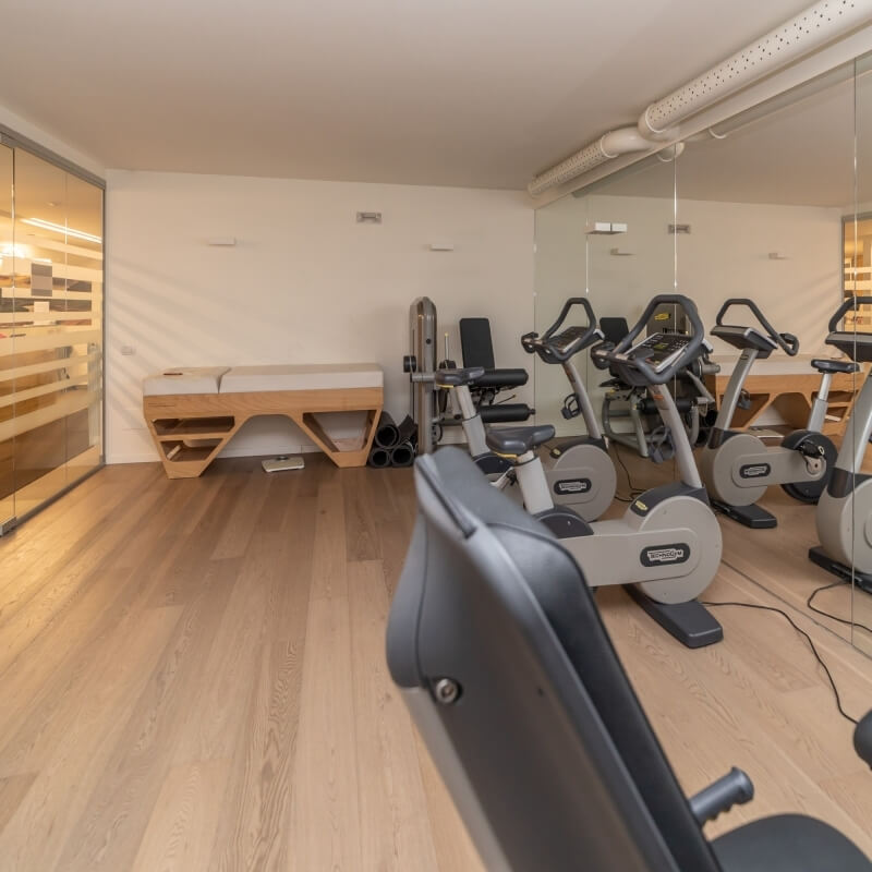 Exercise room with mirrored wall, stationary bikes, wooden bench, and light wood flooring against white walls.
