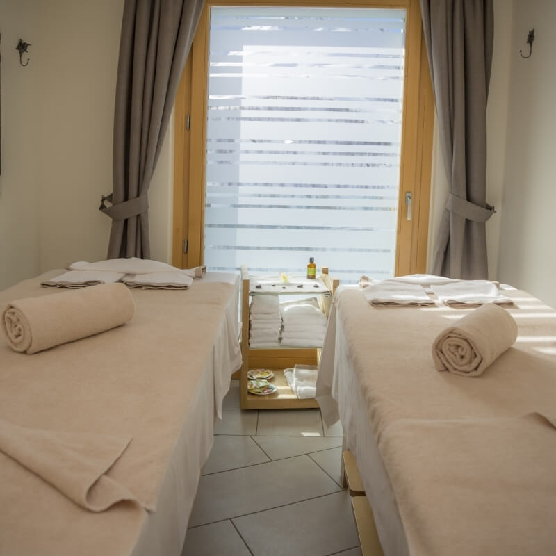 Serene spa room with two massage tables, wooden cart with towels and oils, frosted window, gray curtains, and zigzag tile floor.