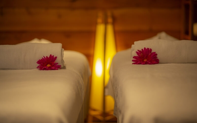 Serene spa room with two beds, white sheets, pink flowers, and warm lighting, creating a calming atmosphere.