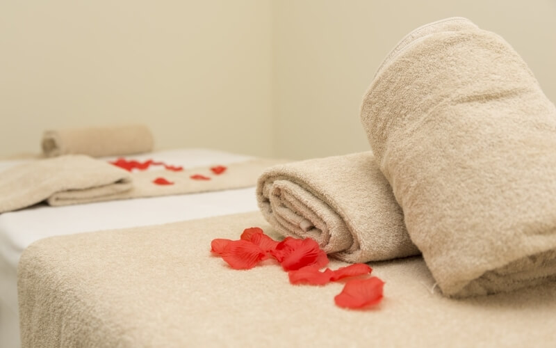 Two light beige towels with sheen on a white table, surrounded by red rose petals against a warm beige wall.